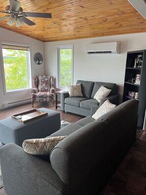 A living room with a ceiling fan, two windows, a grey sofa, a small table, and a bookshelf.