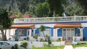A two-story building with a balcony, surrounded by greenery and a mountainous backdrop. - Sea view Apartment P/4 (Benitses, Corfu)