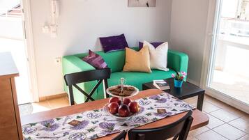 Een eethoek met een tafel gedekt voor twee personen, een groene bank met kussens, een kleine potplant en een raam met uitzicht.