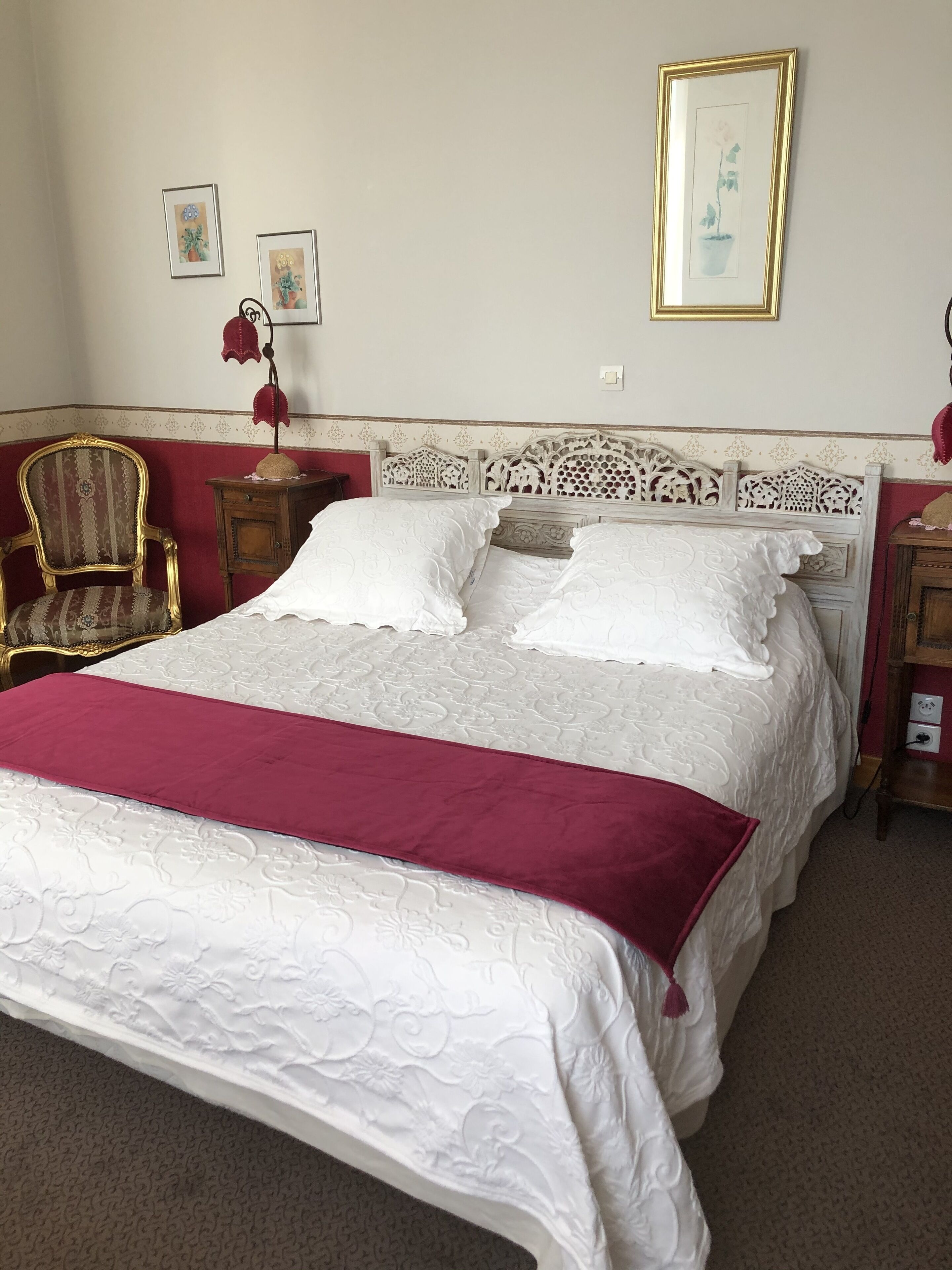 A neatly made bed with a white and red bedspread, a wooden headboard with intricate carvings, and a red chair beside the bed.