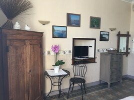 A room with a wooden cabinet, a small table with a potted orchid, two chairs, a television on a wooden stand, a mirror, and a dresser.