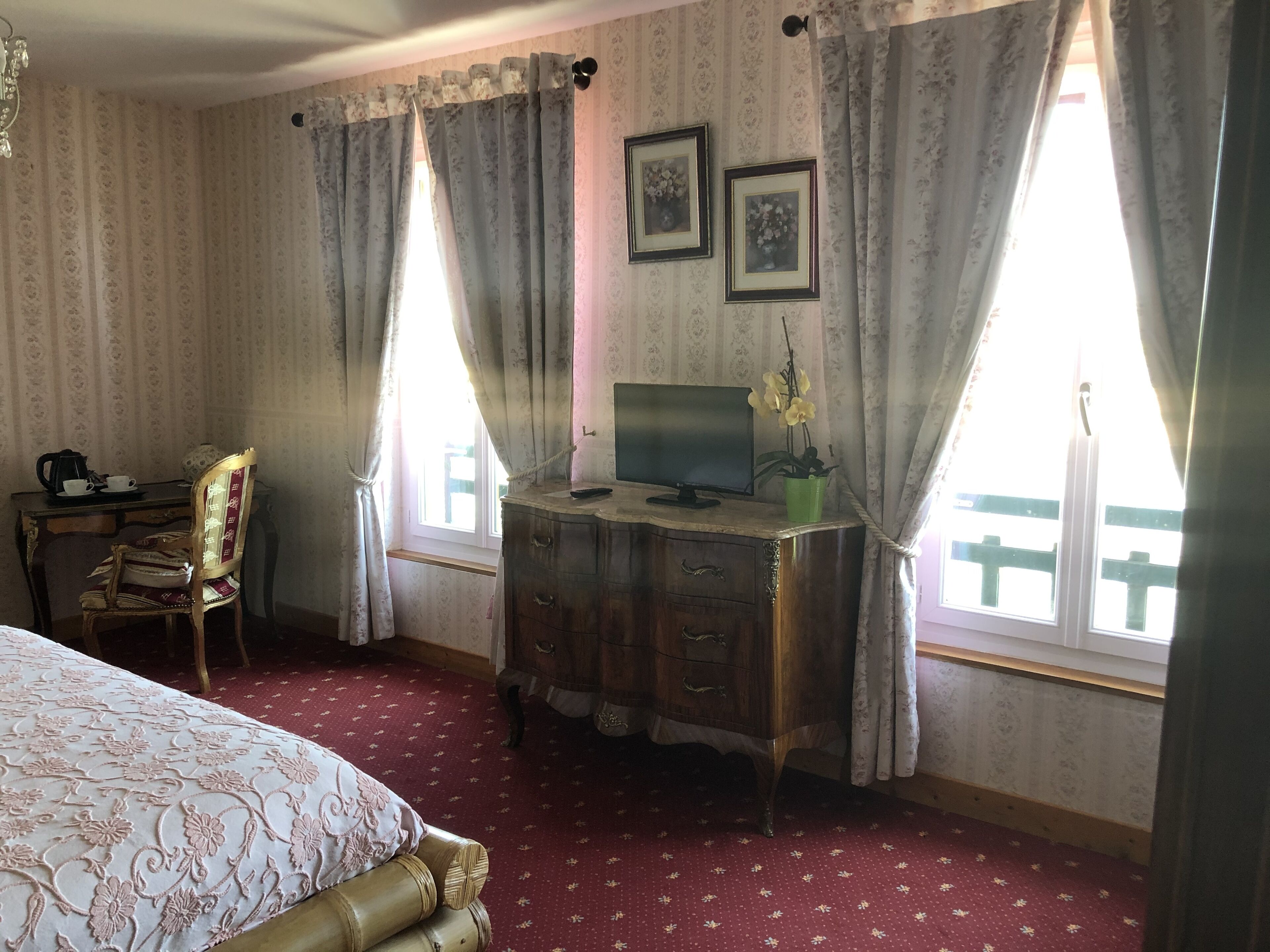 A room with a bed, a wooden dresser, a desk with a computer, and a chair. There are two windows with curtains, and a chandelier hangs from the ceiling.