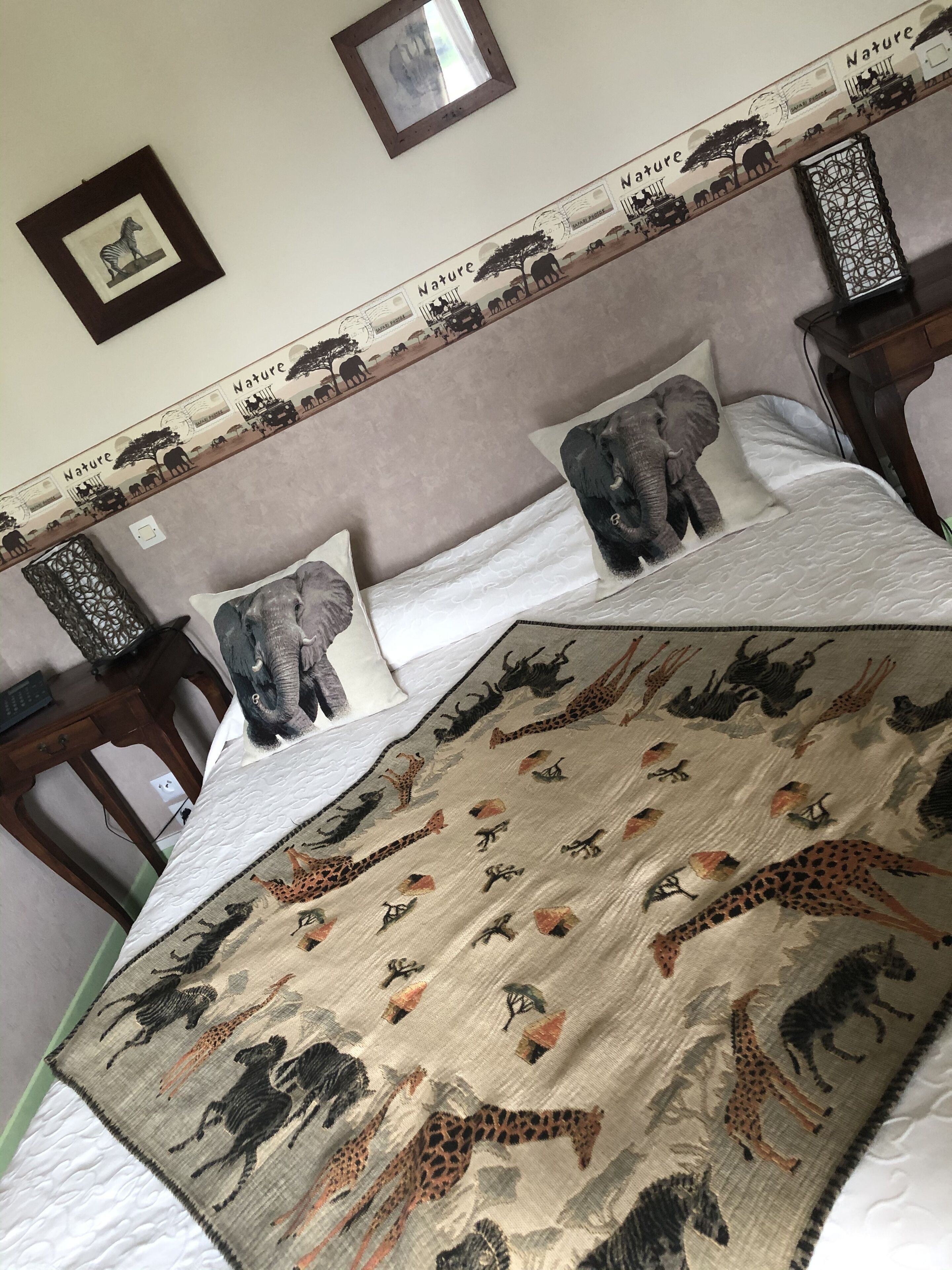 Una cama con colcha de motivos naturales que muestra animales, cabecera con escena natural y un cuadro en la pared.
