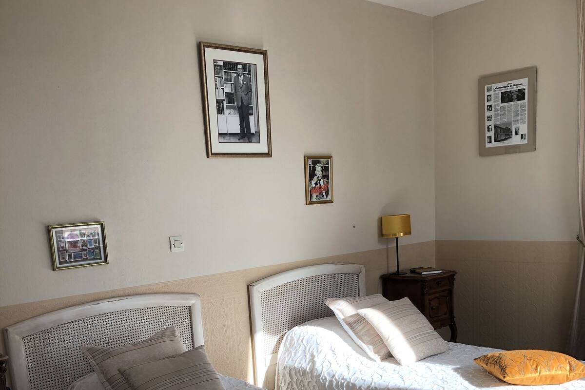 Habitación de hotel con dos camas, cada una con cabecero blanco y colcha estampada. Se observan cuadros enmarcados en las paredes, una lámpara de noche y una mesita con un libro.