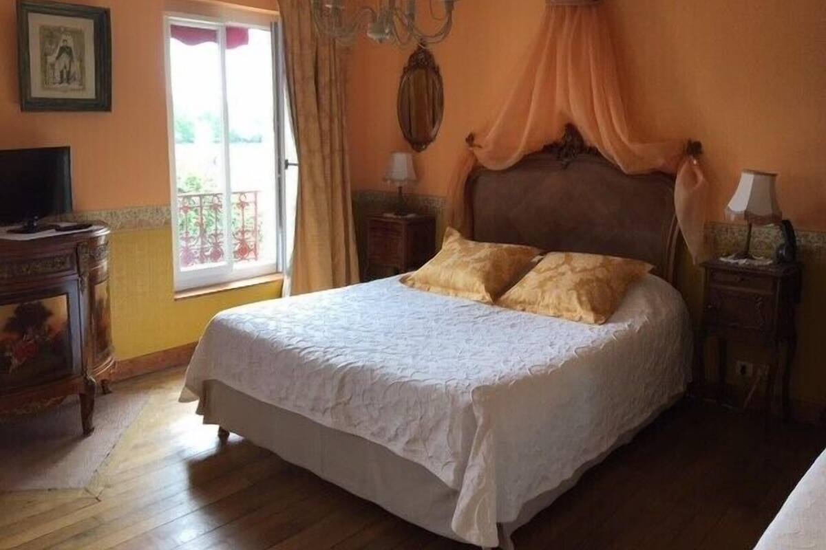 Un dormitorio con una cama grande, un candelabro, una ventana con cortinas y una mesita de noche con lámpara.