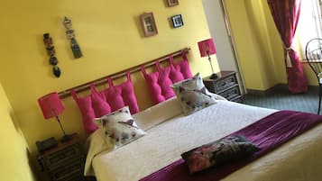 Una cama con una colcha blanca y morada, dos almohadones decorativos, una mesita de noche de madera con una lámpara, y una pared con cuadros y adornos.