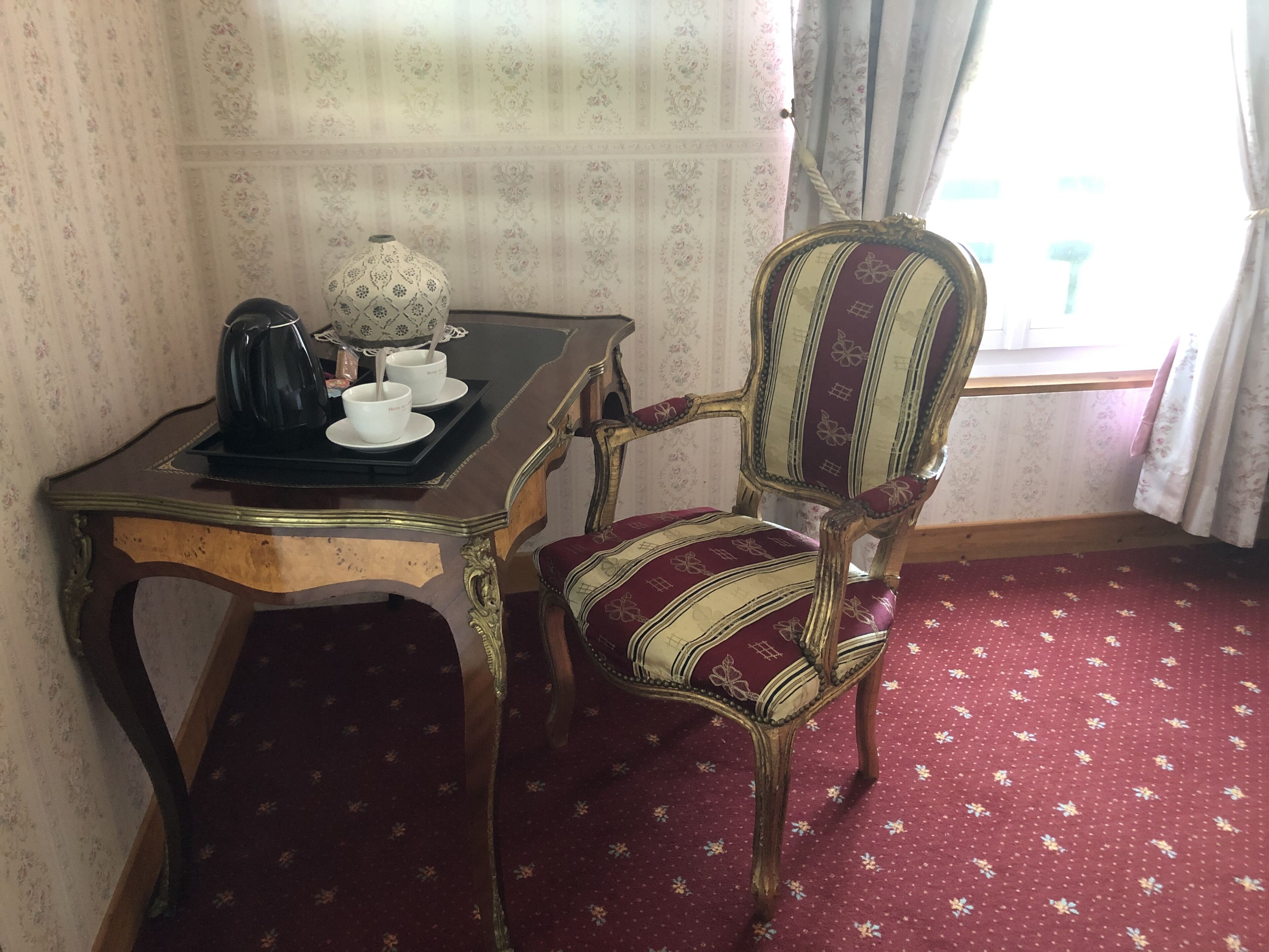 Een kamer met een fauteuil in vintage-stijl, een klein tafeltje met een theepot en kopjes, en een kamerbreed tapijt met dessin.