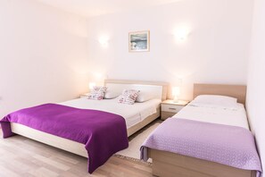 A double bed with a purple blanket, a smaller bed with a purple blanket, a nightstand with a lamp, and a wall-mounted picture.