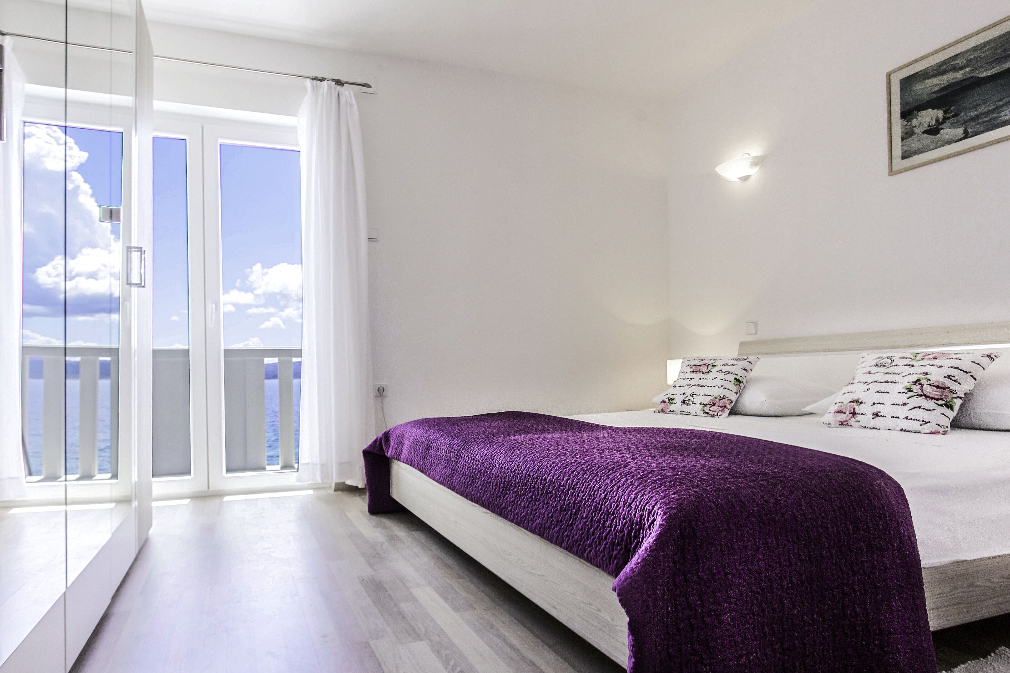 Quarto com cama, paredes brancas, uma luminária no teto e vista através de uma porta de vidro.