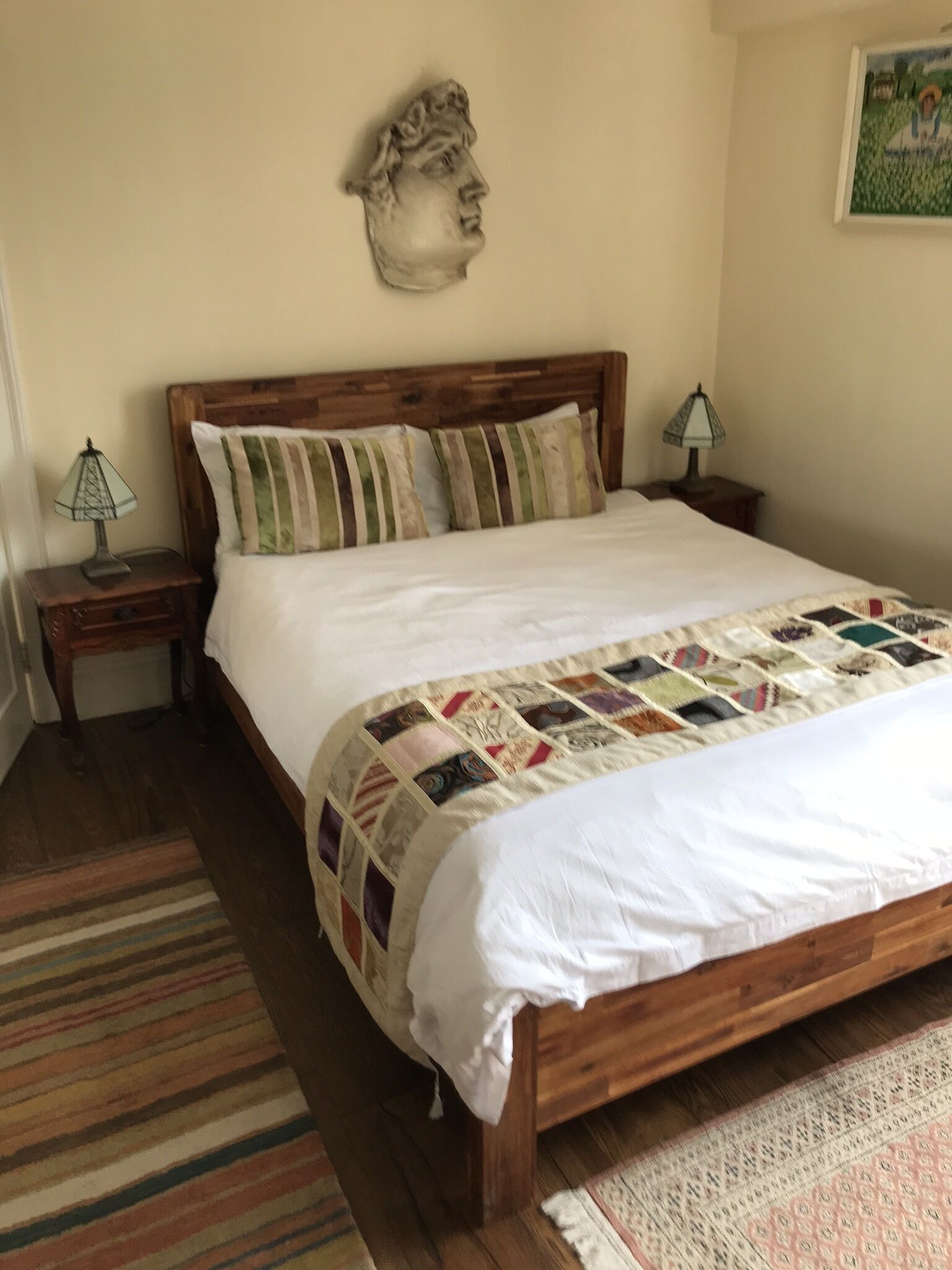 A bedroom with a wooden bed, striped pillows, a quilt, two bedside tables with lamps, a wall-mounted headboard, and a rug.
