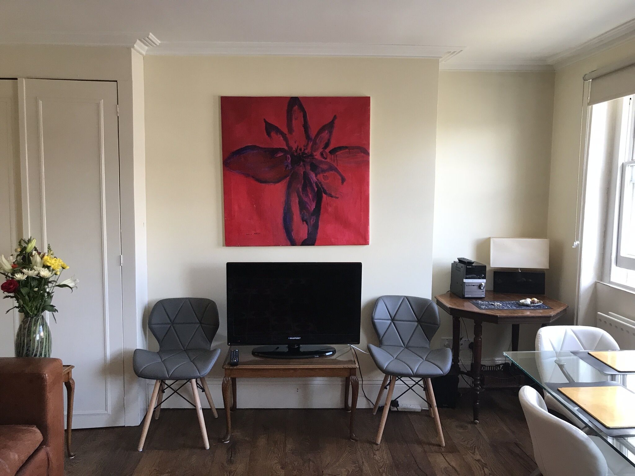 Een moderne woonkamer met een flatscreen-tv, twee grijze fauteuils, een houten salontafel, een abstract schilderij in rood, een vaas met bloemen, en een tafeltje met een lamp en een printer.