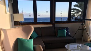 A living room with a sofa, armchair, and a glass coffee table. There are windows with a view of the sea and palm trees. - Apartment PANORAMA Great View, WiFi, 50m beach (Costa Calma)