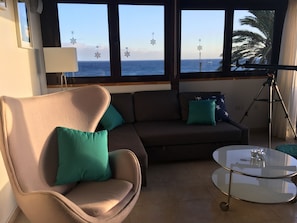 A living room with a sofa, armchair, and a glass coffee table. There are windows with a view of the sea and palm trees.