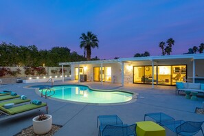 A modern outdoor pool area with lounge chairs, a covered seating area, and a well-lit building. - Lena's Hideaway – Swanky Mid-Mod Palm Springs Retreat (Palm Springs)