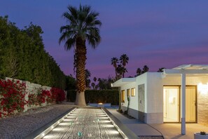 A modern building with a pathway leading to a door, surrounded by palm trees and red flowers.