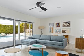Un salon moderne avec un canapé d’angle, une table basse ronde, un ventilateur de plafond et une grande fenêtre donnant sur une piscine.