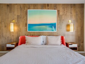 A bedroom with a large bed, wooden wall paneling, a painting of a beach, and two bedside lamps. - Lena's Hideaway – Swanky Mid-Mod Palm Springs Retreat (Palm Springs)