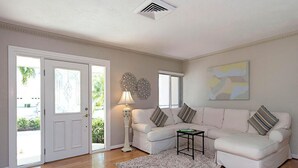 A living room with a white sofa, a small side table, and a lamp. There is a door with glass panels and a view of greenery outside. - 3-bedroom, 2- bath home with wide water views. (Marco Island)