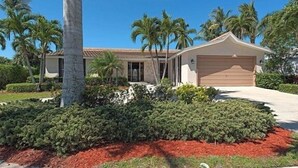 A single-story house with a beige garage door, surrounded by a well-maintained garden with red mulch and various shrubs. - 3-bedroom, 2- bath home with wide water views. (Marco Island)