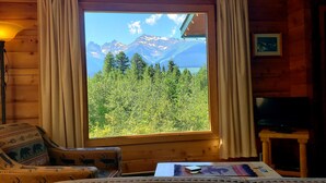 Un cuarto acogedor con un ventanal grande que ofrece vistas a la montaña, una lámpara de pie de madera, un sillón estampado y una mesita con un control remoto.