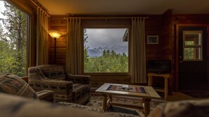 Interior acogedor de una cabaña con un amplio ventanal que ofrece vistas a la montaña, un sofá mullido, una mesa de centro de madera y una lámpara.