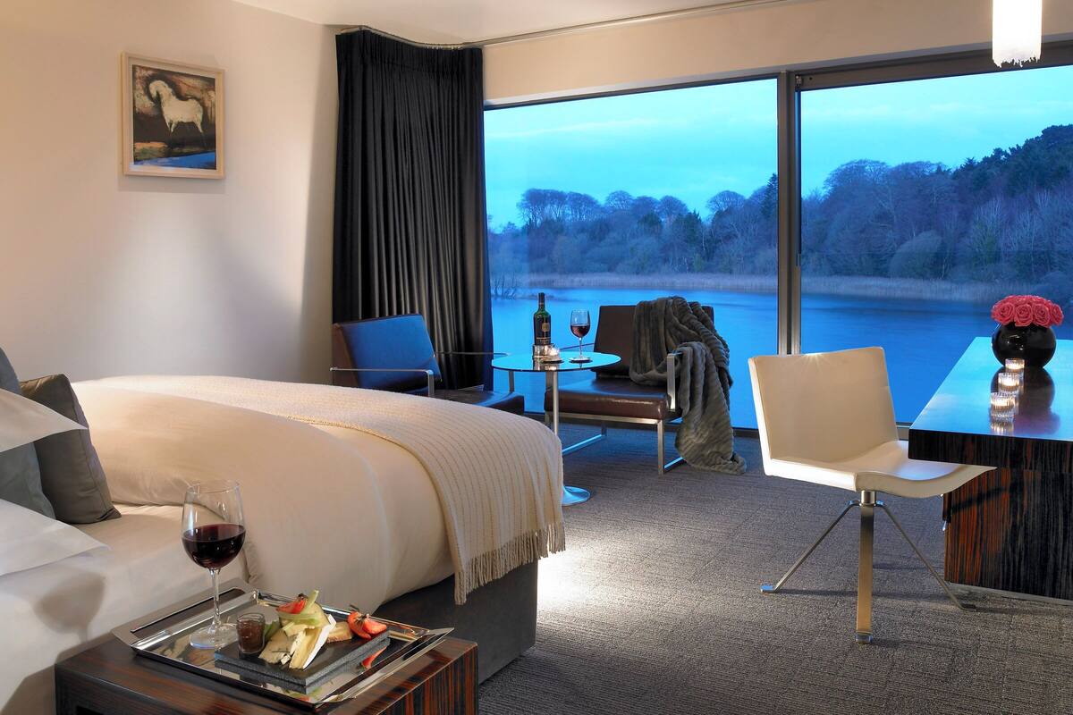 A hotel room with a large bed, a dining table with a vase of flowers, and a view of a lake.