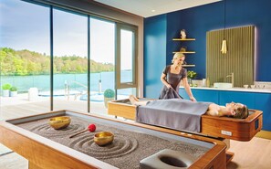 Una stanza per massaggi con vista su un lago, un lettino da massaggio e un’area relax con campane tibetane.