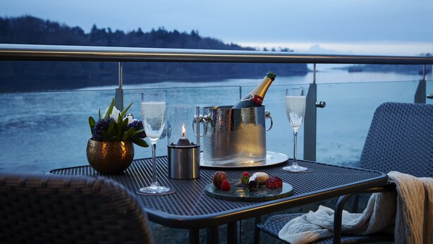 A table set with a bottle of champagne, glasses, a candle, and a plate of pastries on a balcony overlooking a body of water. - Ice House Hotel (Ballina)