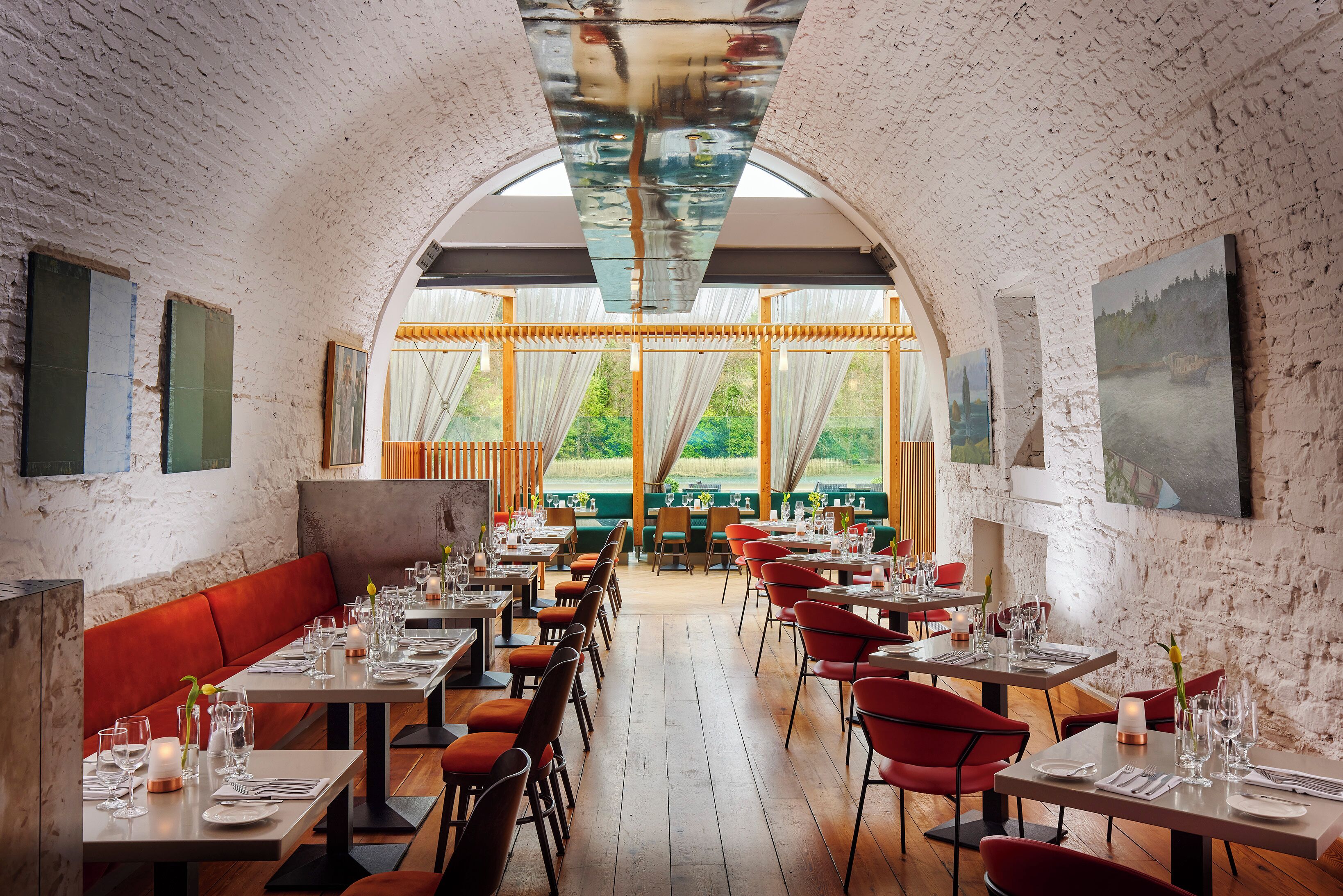 A restaurant with a rustic brick wall, wooden flooring, and red upholstered seating. Tables are set with white tablecloths and candles.