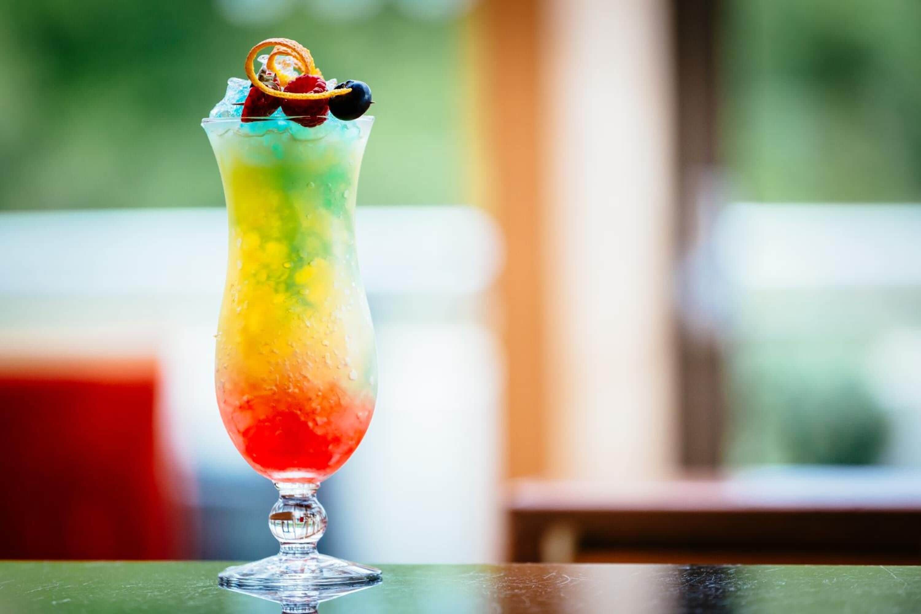 A colorful layered drink in a tall glass with a garnish, placed on a table with a blurred background.