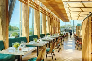 A restaurant with long wooden tables, green upholstered booths, and large windows offering a view of the outdoors. - Ice House Hotel (Ballina)