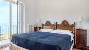 Un dormitorio con una cama grande, un cabecero de madera, dos mesitas de noche con lámparas, un radiador y un balcón con vistas.