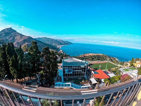 A coastal hotel with a prominent blue building, a road with a pedestrian crossing, and a view of the sea and mountains.