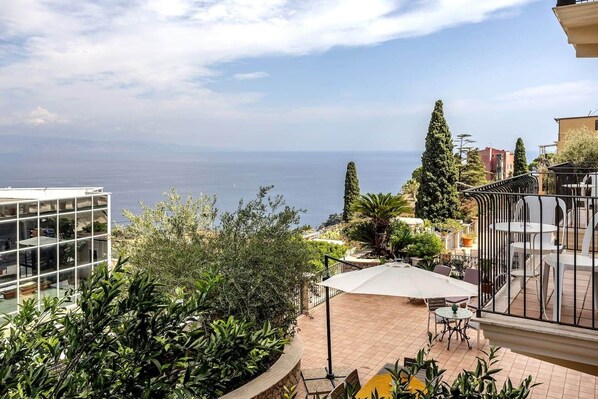 A terrace with outdoor seating, a view of the sea, and surrounding greenery.