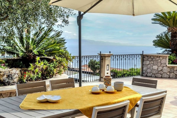 An outdoor patio with a table set for dining, surrounded by chairs, and a view of the sea.