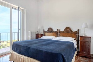 A bedroom with a large bed, a wooden headboard, two bedside tables with lamps, a radiator, and a balcony with a view.