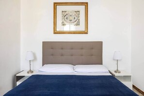 Ein ordentlich bezogenes Bett mit blauer Decke, zwei Nachttischlampen und einem gerahmten Bild darüber.