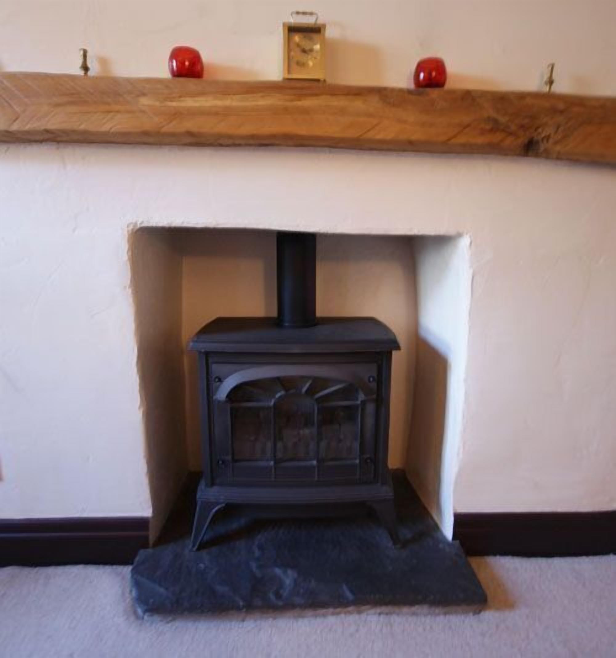 A black wood-burning stove with a glass door, positioned under a wooden mantelpiece with red apples and a clock.
