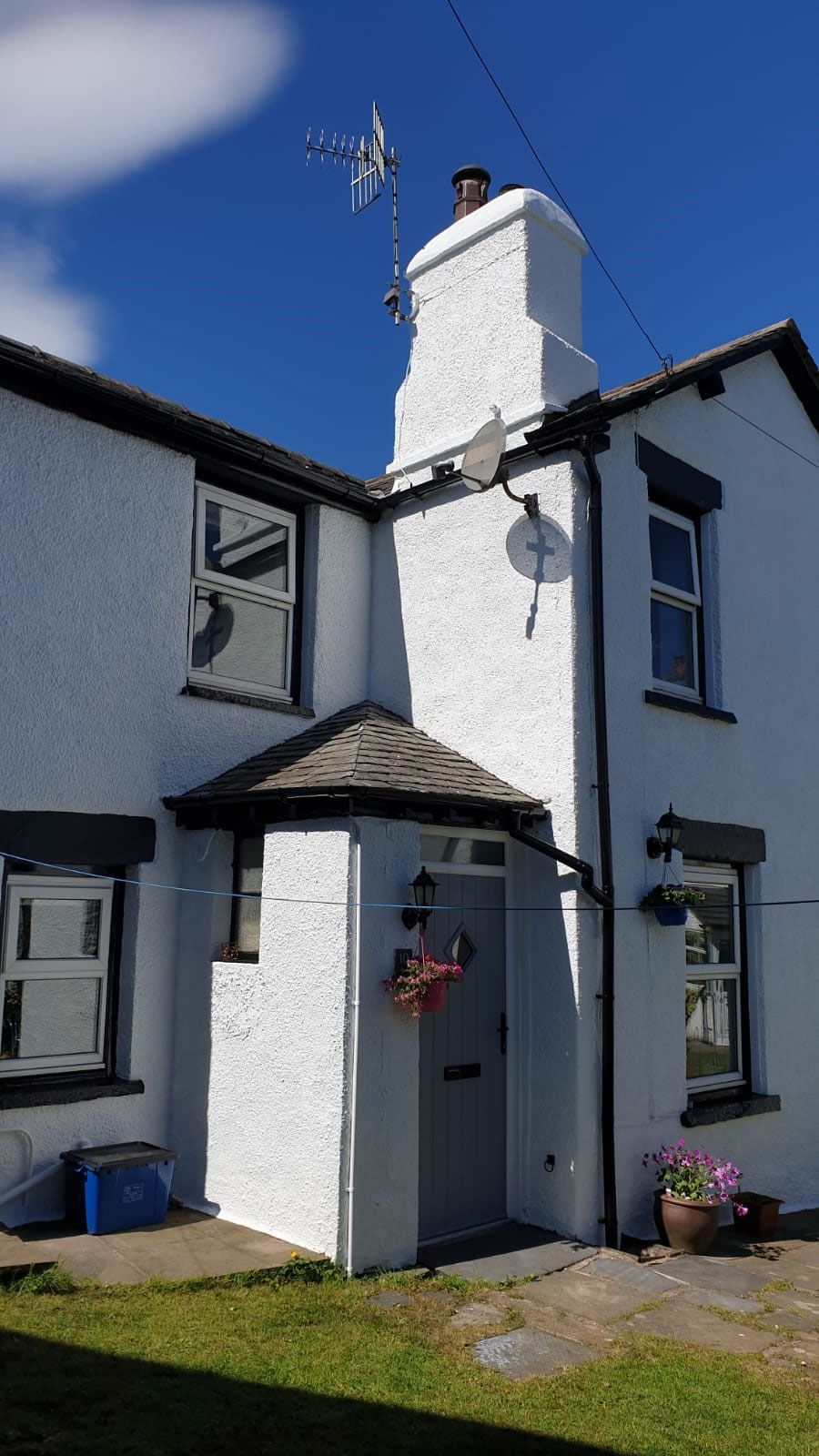 Un bâtiment blanc avec une cheminée, une antenne parabolique et un petit porche agrémenté d’un pot de fleurs.