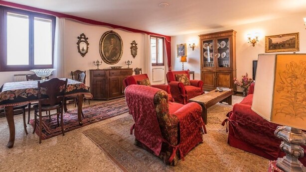 A traditional living room with a dining area, featuring red upholstered furniture, a wooden cabinet with glass doors, and a large framed portrait on the wall.