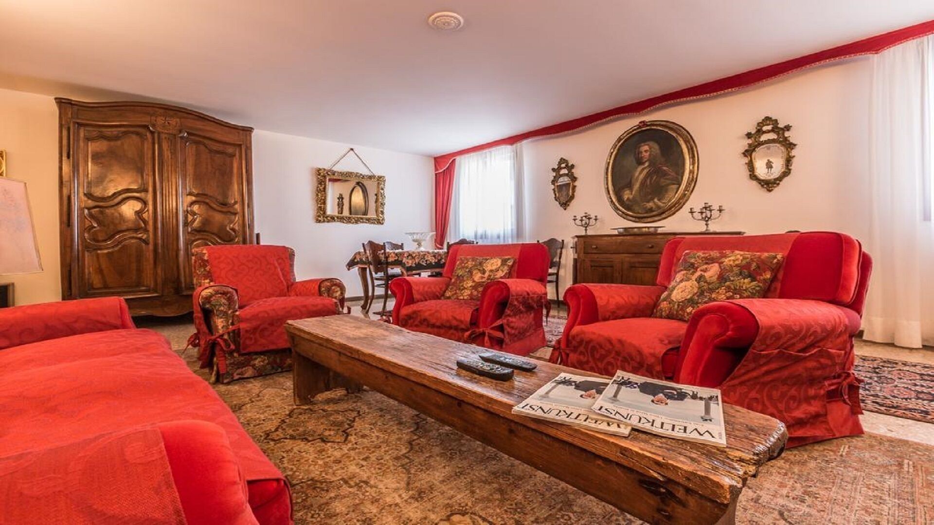 Un salon traditionnel avec des meubles rembourrés de couleur rouge, une table basse en bois sur laquelle reposent des journaux, et une décoration classique comprenant un grand miroir et des œuvres d’art encadrées accrochées aux murs.