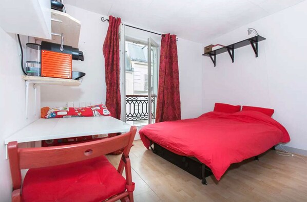 A small, well-lit room with a single bed, a table with a chair, and a shelf with a red object.