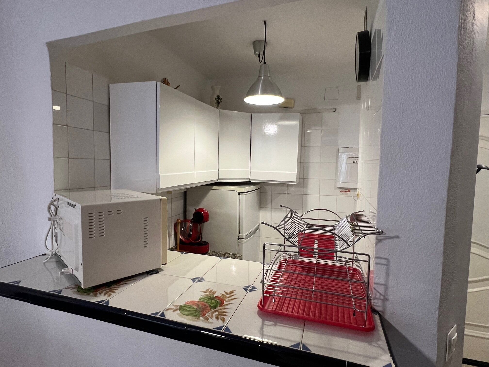 A compact kitchen with white cabinets, a microwave, a red drying rack, and a hanging light fixture.
