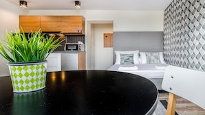 Habitació d'hotel moderna amb taula de dinar negra, una planta en test, llits amb coixins estampats i una cuina al fons.