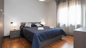 Una cama bien tendida con un edredón azul oscuro, dos almohadas y una lámpara de noche sobre una mesita de madera.