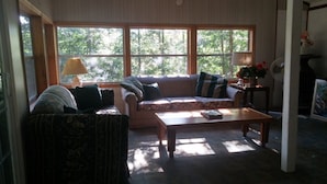 A living room with a sofa, armchair, and coffee table. There are two windows with a view of trees. A lamp and a vase with flowers are on a side table.