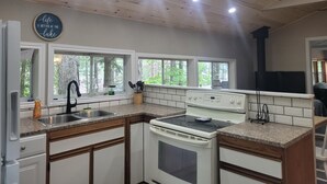 A kitchen with white cabinets, a stove, and a sink. There are windows with a view of trees outside.
