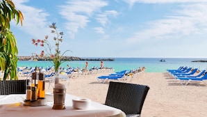 Un espace de restauration aménagé au bord de la plage, avec une table dressée pour deux, donnant sur une plage de sable fin, agrémentée de chaises longues bleues et offrant une vue dégagée sur la mer.