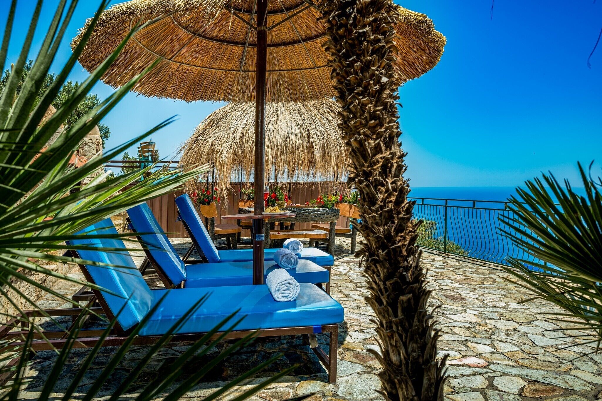 Un área de descanso en la playa con sillones azules, una sombrilla de paja y vista al mar.