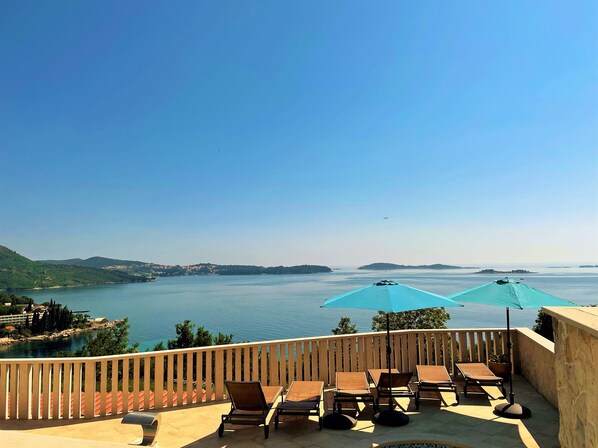 Un balcon doté de chaises longues et de parasols, offrant une vue sur une mer calme et des îles lointaines.
